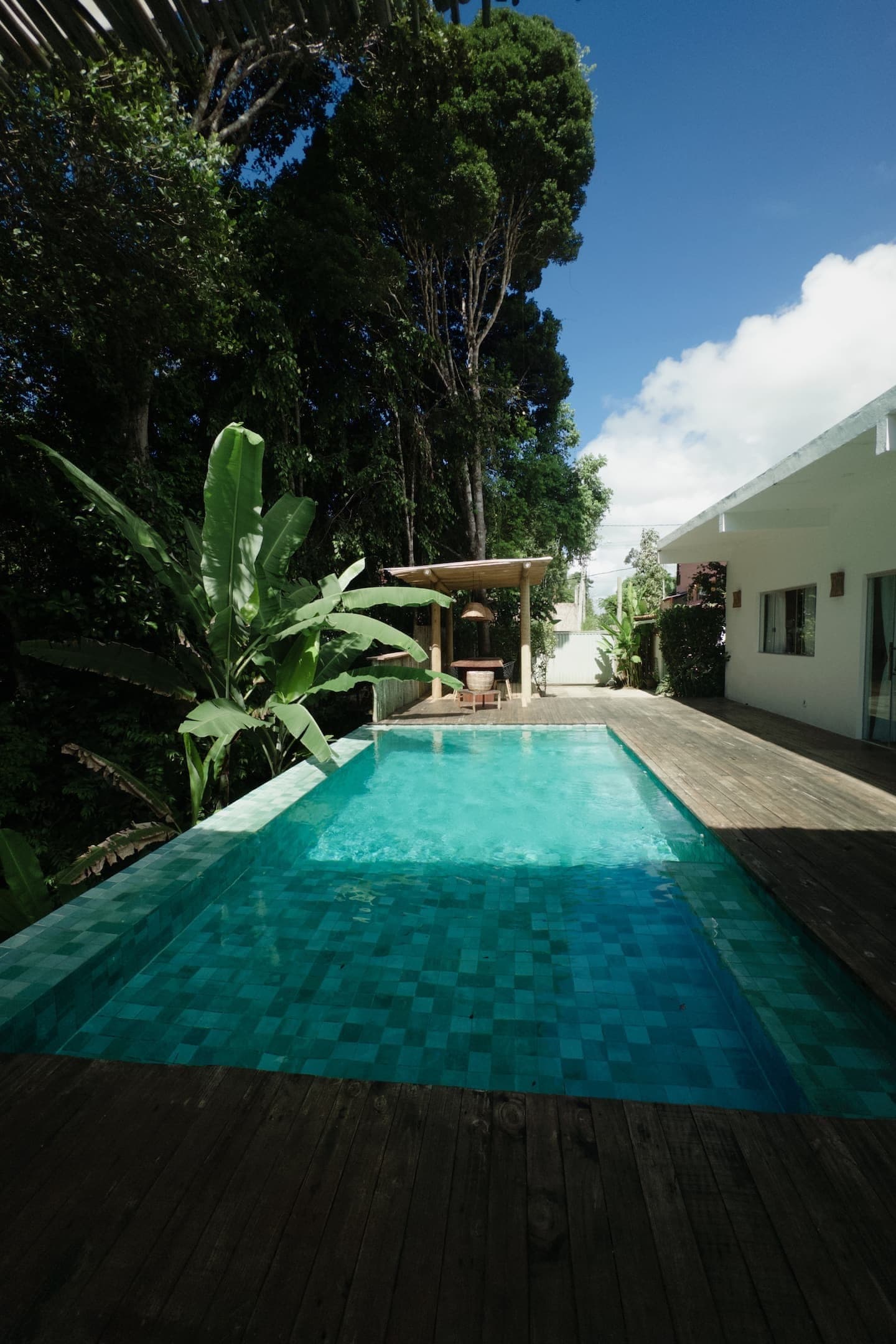 Piscina comprida entre bananeiras e deck de madeira, com a casa ao fundo